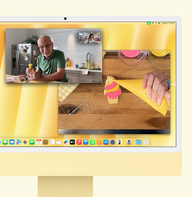iMac i gul. Demonstrasjon av Bordvisning. Skjermen viser en baker i en videosamtale med en annen person. Én skjerm viser bakeren og mannens ansikt. Den andre skjermen viser bakerens hender, som tar glasur på en kjeks