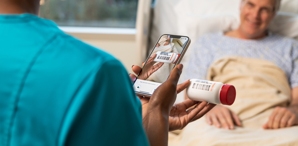 En sygeplejerske i blågrøn uniform scanner en patients medicin med sin iPhone.