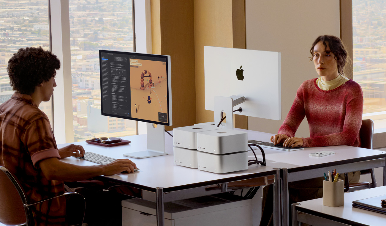 Two people sitting at two desks facing each other, four Mac Studios are on one desk in two stacks of two, two Studio Displays are on the desks, one screen shows a various project in-process in animation and editing apps, the other shows the back side of the display